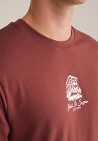 Bordeaux bomulds T-shirt med rund hals. Har et hvidt grafisk design af en bygning og teksten "Hotel Il Sogno EST. 1998" på fronten.