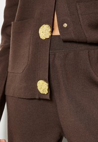 Braune Strickjacke mit zwei großen goldfarbenen Texturknöpfen und einer aufgesetzten Tasche. Koordinierende braune Hose am unteren Rand sichtbar.