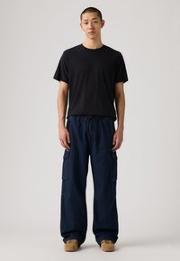 Μαύρο κοντομάνικο μπλουζάκι με στρογγυλή λαιμόκοψη, σε συνδυασμό με φαρδιά σκούρα μπλε παντελόνια καμπαρντίνας με πλαϊνές τσέπες. Φορεμένο με ταμπά αθλητικά παπούτσια. Απλός σχεδιασμός.