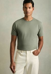 T-shirt verde oliva a maniche corte realizzato in morbido tessuto, abbinato a jeans in denim color crema. Il modello si trova in piedi con una mano in tasca su uno sfondo neutro.