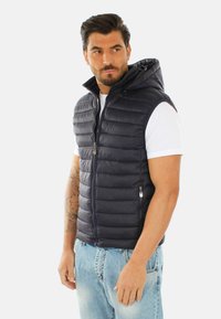 Gilet imbottito blu navy con zip frontale e cappuccio, caratterizzato da cuciture orizzontali e tasche laterali, indossato sopra una t-shirt bianca e jeans blu.