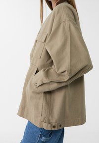 Beige Canvas-Jacke mit lockerem Schnitt und großen Seitentaschen. Mit braunem Kragen und Knopfakzenten an den Ärmeln und dem Saum.