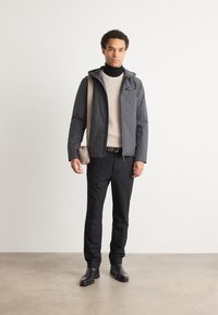 Giacca grigia con cappuccio sopra un maglione leggero, pantaloni neri e stivali neri; porta una piccola borsa beige; texture lisce e design informale.
