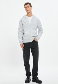 Sudadera gris con cremallera y capucha ajustable, combinada con jeans negros y zapatos gruesos negros. La tela parece suave con una textura lisa.