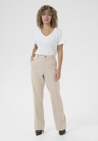 Beige maßgeschneiderte Hose mit einem strukturierten Streifenmuster und einem Knopfverschluss vorne, kombiniert mit einem schlichten weißen V-Ausschnitt-T-Shirt und schwarzen High Heels.