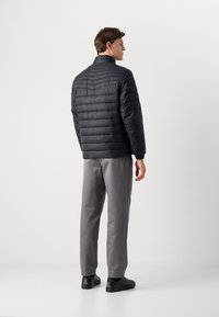 Young man standing facing right, wearing a black quilted jacket, gray pants, and black sneakers against a plain light background.