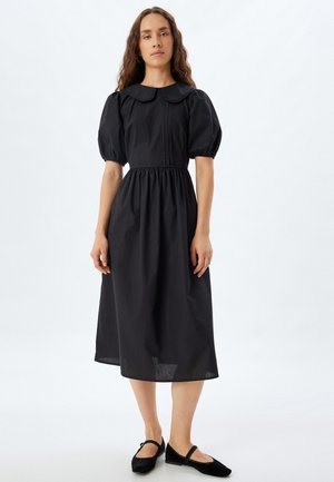 Schwarzes Kleid mit puffärmel, runder Kragen, tailliert und knielang. Glattes, strukturiertes Material; kombiniert mit schwarzen Schuhen.