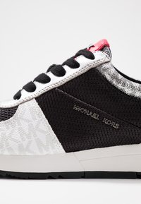 Svart och vit sneaker med nät- och lädermaterial, med ett mönstrat sidor, tjocka svarta snören och en metallisk logotypdetalj.