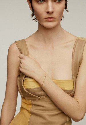 Top de cuero marrón con acentos amarillos que presenta un escote cuadrado y tirantes anchos. La modelo lleva una pulsera delicada con adornos de colores.