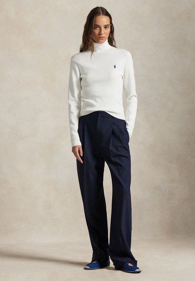 Mujer de pie con un suéter de cuello alto blanco, pantalones holgados azul marino y zapatos azules sin cordones sobre un fondo neutro.
