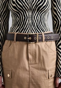Bruine faux lederen riem met gouden hardware, gedragen over een beige rok met voorzakken, aangevuld met een zwart-wit gestreepte top.