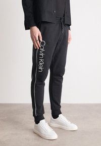 Zwarte joggerbroek met het witte Calvin Klein-logo langs het linkerpand, voorzien van een trekkoord in de taille en taps toelopende enkels. Draagt met witte sneakers.