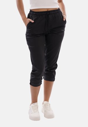Persona che indossa pantaloni neri corti con risvolti e sneakers bianche, mani in tasca, in piedi davanti a uno sfondo bianco.