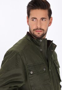 Giacca verde oliva con chiusura a zip e bottoni a pressione, due tasche sul petto, spalle strutturate e collo in corduroy marrone. Texture opaca.