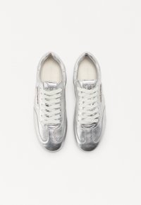 Baskets métalliques argentées avec des lacets blancs, présentant une texture lisse, une forme arrondie et un logo sur la languette et sur le côté.