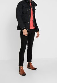 Černá vatovaná bunda se zapínáním na patentky, dvě přední kapsy, nošená přes korálový svetr, v kombinaci s černými úzkými kalhotami a hnědými chelsea botami.