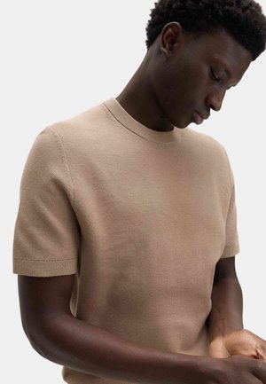 Junger Mann mit dunkler Haut und kurzen lockigen Haaren, der nach unten schaut, trägt einen beigen Strickpullover mit kurzen Ärmeln vor einem schlichten hellen Hintergrund.