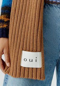 Brauner, gerippter Strickschal mit einem rechteckigen weißen Etikett, auf dem der Text "oui" in Schwarz steht, gehalten vor Denim-Stoff.