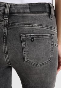 Jeans de mezclilla gris oscuro con un bolsillo trasero que cuenta con un cierre de botón a presión plateado y una parche de cuero negro en la cinturilla.