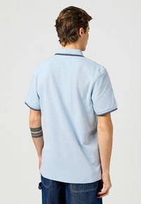 Ljusblå polo med mjuk textur, korta ärmar och kontrasterande mörkblå krage och ärmslut, visad från ryggen.