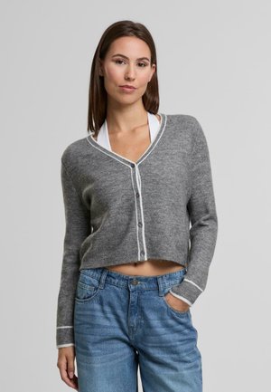 Cárdigan gris corto hecho de material suave, con un diseño de escote en V, borde blanco en los extremos y botones en el frente. Lleva puesto un top blanco.