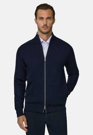 Homme portant un pull zippé bleu marine sur une chemise blanche à col, debout avec une main dans la poche devant un fond uni.