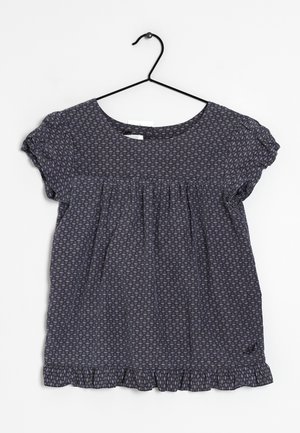 Top de manga corta en azul oscuro con un dobladillo con volantes, que presenta un patrón geométrico de pequeños triángulos en tonos más claros. Material de algodón, ajuste holgado.