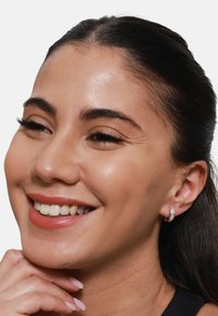 Orecchini a cerchio argentati con una superficie texturizzata, indossati da un modello con capelli scuri e lisci, che sorride e appoggia il mento su una mano.