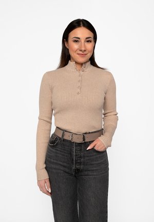 Giovane donna con lunghi capelli scuri che indossa un maglione beige a costine con bottoni, jeans neri a vita alta e cintura beige, in piedi davanti a uno sfondo bianco.