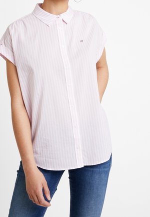 Kobieta ubrana w różową koszulę w paski na krótki rękaw i niebieskie dżinsy, stojąca z jedną ręką w kieszeni.