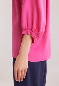 Blusa fucsia con maniche a sbuffo, caratterizzata da dettagli a bottone al polsino. Realizzata in tessuto morbido, ha una vestibilità comoda e orlo dritto.