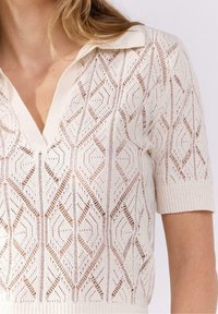Stickad polo-liknande topp i off-white med ett diamantmönster, som har korta ärmar och en strukturerad, andningsbar design.