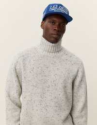 Hellgrauer gestrickter Rollkragenpullover mit einer gesprenkelten Textur, kombiniert mit einer blauen Mütze, die weiße Schriftzüge und Logos enthält.