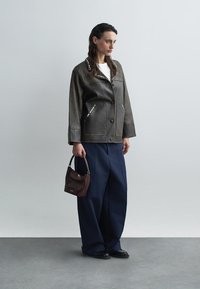 Smeđa kožna jakna sa srebrnim zakovicama, uparena s mornarskoplavim hlačama širokih nogavica i malom torbicom u boji burgundca. Model stoji nasuprot neutralnoj pozadini.