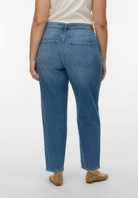 Vrouw die blauwe, high-waist, rechte jeans en beige platte schoenen draagt, staand tegen een effen witte achtergrond, met haar rug naar de camera gericht.