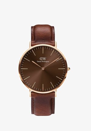 Pruun nahast rihmaga käekell ümmarguse pruuni kellapaneeli, kuldsete tunni markerite ja kätega. Omab minimalistlikku disaini koos brändi logoga.