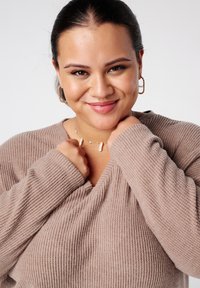 Pull à manches longues, en ribbed, avec un col en V de couleur beige douce. Le modèle porte un collier délicat et des boucles d'oreilles créoles, souriant doucement.