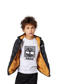 Gutt med krøllet hår iført åpen svart dunjakke med oransje fôr og en lys grå Timberland t-skjorte med logo på fronten.