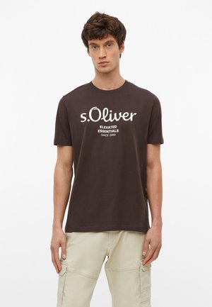 Młody mężczyzna w brązowej koszulce z krótkim rękawem z napisem „s.Oliver Elevated Essentials Since 1969” oraz beżowych spodniach typu cargo, stojący na białym tle.