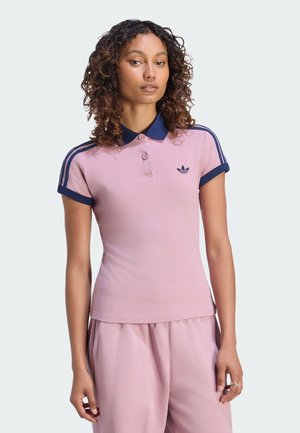 Kobieta o kręconych włosach, ubrana w jasnoróżową koszulkę polo Adidas z ciemnoniebieskim kołnierzykiem i paskami, w zestawie z pasującymi różowymi spodniami.