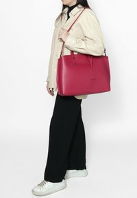 Rode leren draagtas met dubbele handvatten en een label, met een gladde textuur en gestructureerd ontwerp, gedragen door een persoon in een lichte jas.