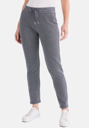 Frau trägt eng anliegende, strukturierte graue Hose mit Kordelzug, kombiniert mit weißen Sneakers, steht vor einem schlichten Hintergrund.