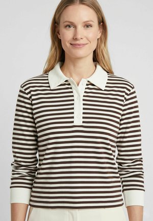 Femme aux longs cheveux blonds portant un polo à manches longues rayé horizontalement marron et blanc avec un col blanc.