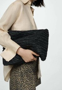 Borsa a tracolla nera intrecciata con un design testurizzato, tenuta sotto il braccio. La modella indossa una camicia beige chiaro e una gonna con stampa leopardo.
