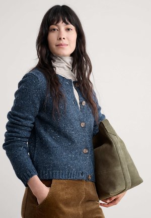 Gilet tricoté à pois navy avec des boutons, superposé à un col roulé léger. Pantalons en velours côtelé marron, tenant un sac en daim vert. Fond neutre.