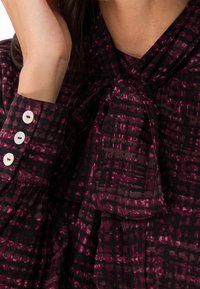 Blouse à manches longues en bordeaux foncé avec un motif texturé, dotée d'un col à nœud et d'un poignet boutonné avec des boutons blancs.