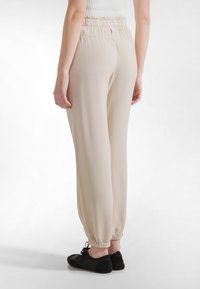 Beige, lette stofbukser med elastisk talje, tilspidsede ben og samlede manchetter. Bæres med sort fodtøj.