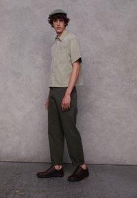 Korte mouwen, olijfgroen overhemd met knopen, donkergroene relaxte broek en donkerbruine veterschoenen met minimale afwerking.