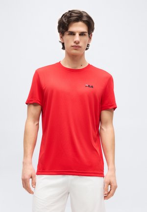 Noor mees, kes kannab punast lühikeste varrukatega FILA särki ja valgeid pükse, seisab ühevärvilise heleda tausta ees.