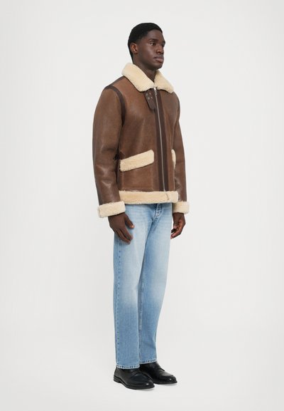 Braune Shearling-Jacke mit cremefarbenen Akzenten, Reißverschluss, zwei Vordertaschen und passenden Shearling-Bündchen, kombiniert mit hellblauen Jeans und schwarzen Schuhen.